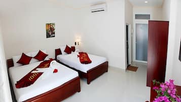 A hotel room with two beds, each with red pillows and a red towel. There is a small painting on the wall, a door to the right, and a wooden cabinet with a vase of flowers.