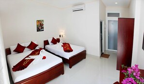 A hotel room with two beds, each with red pillows and a red towel. There is a small painting on the wall, a door to the right, and a wooden cabinet with a vase of flowers.