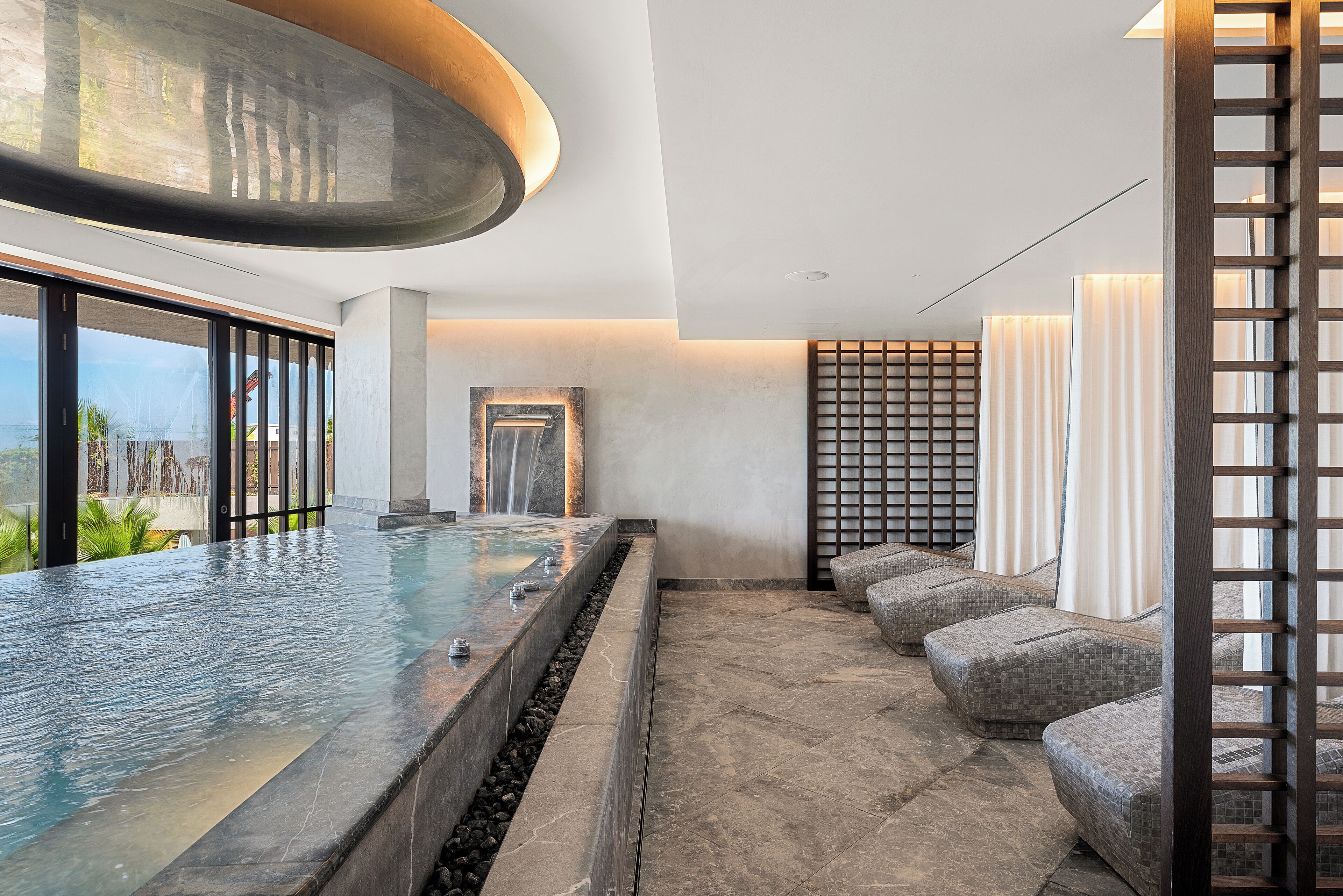 A modern indoor pool area with a long rectangular pool, stone seating, and a view of the outdoors.