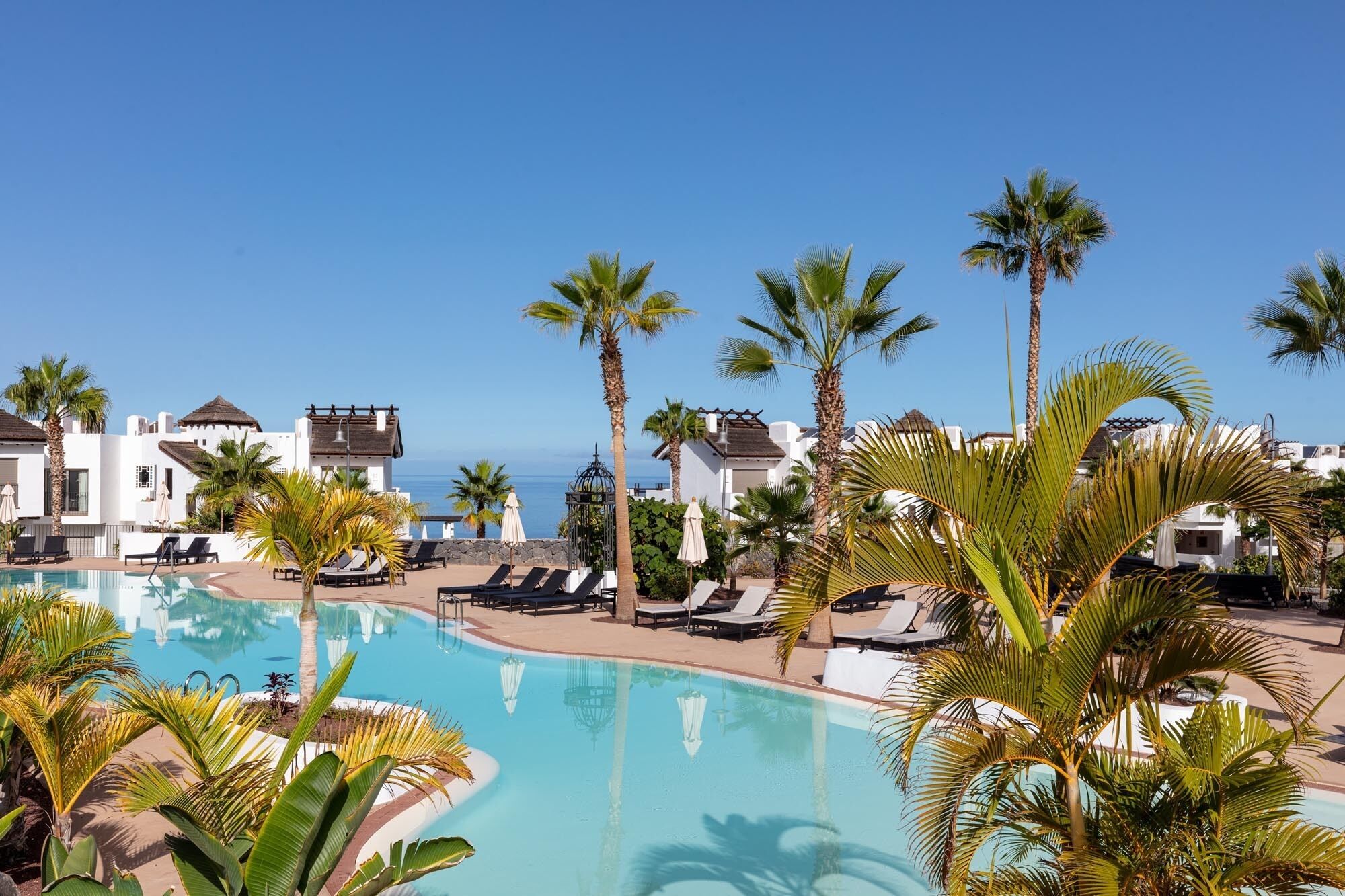 A resort with a swimming pool, palm trees, and white buildings with red-tiled roofs.