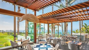 Een ruim buitenterras met meerdere tafels en stoelen, grote ramen met uitzicht op een golfbaan en palmbomen, en een houten pergola.