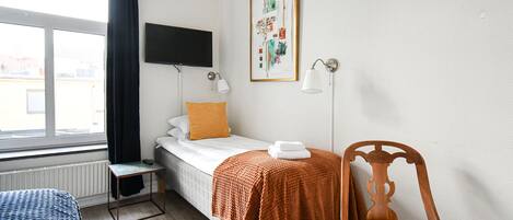 Un dormitorio pequeño y ordenado con una cama individual, una silla, una mesa y un televisor montado en la pared.