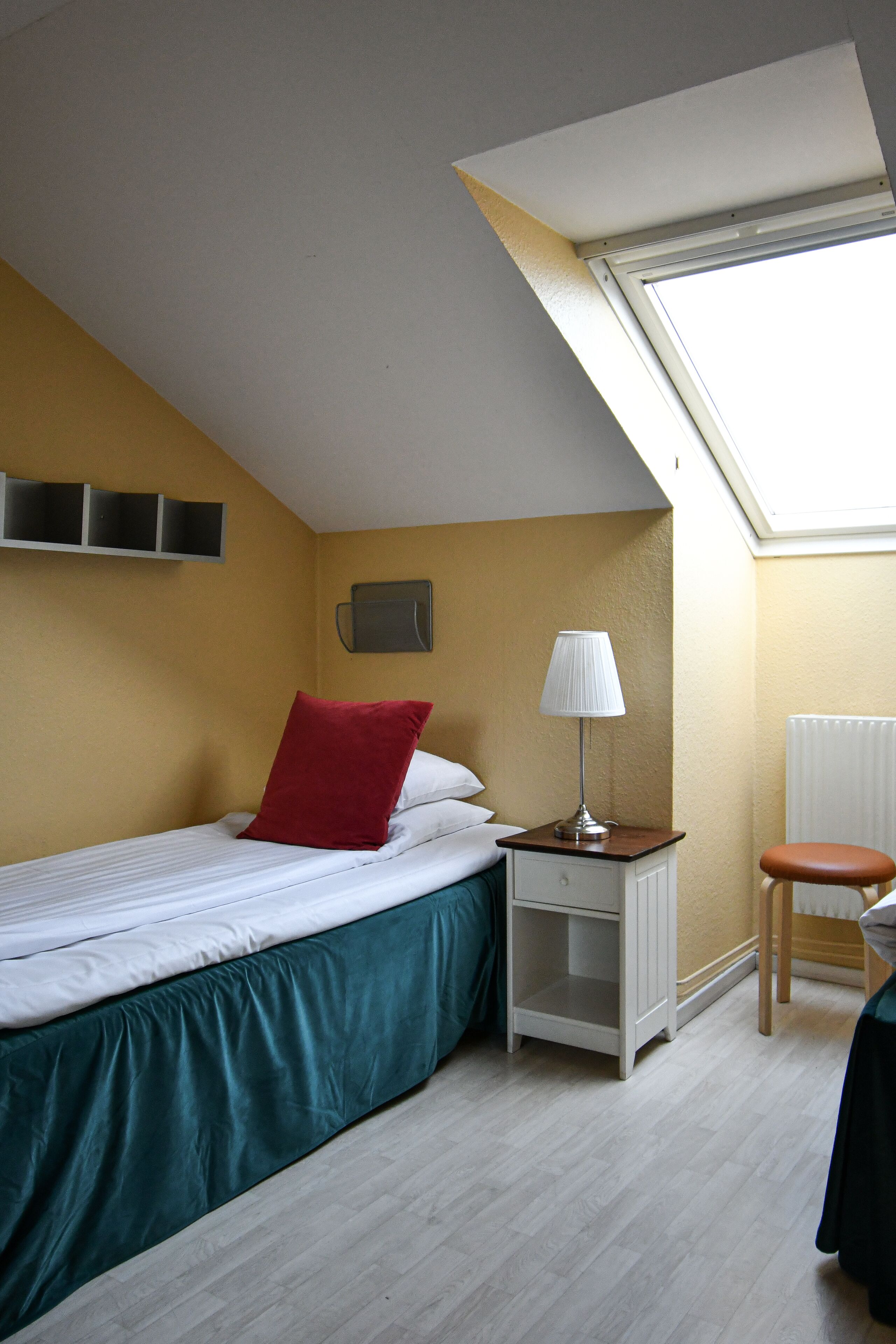 A small, well-lit room with a single bed, a nightstand, a chair, and a skylight.