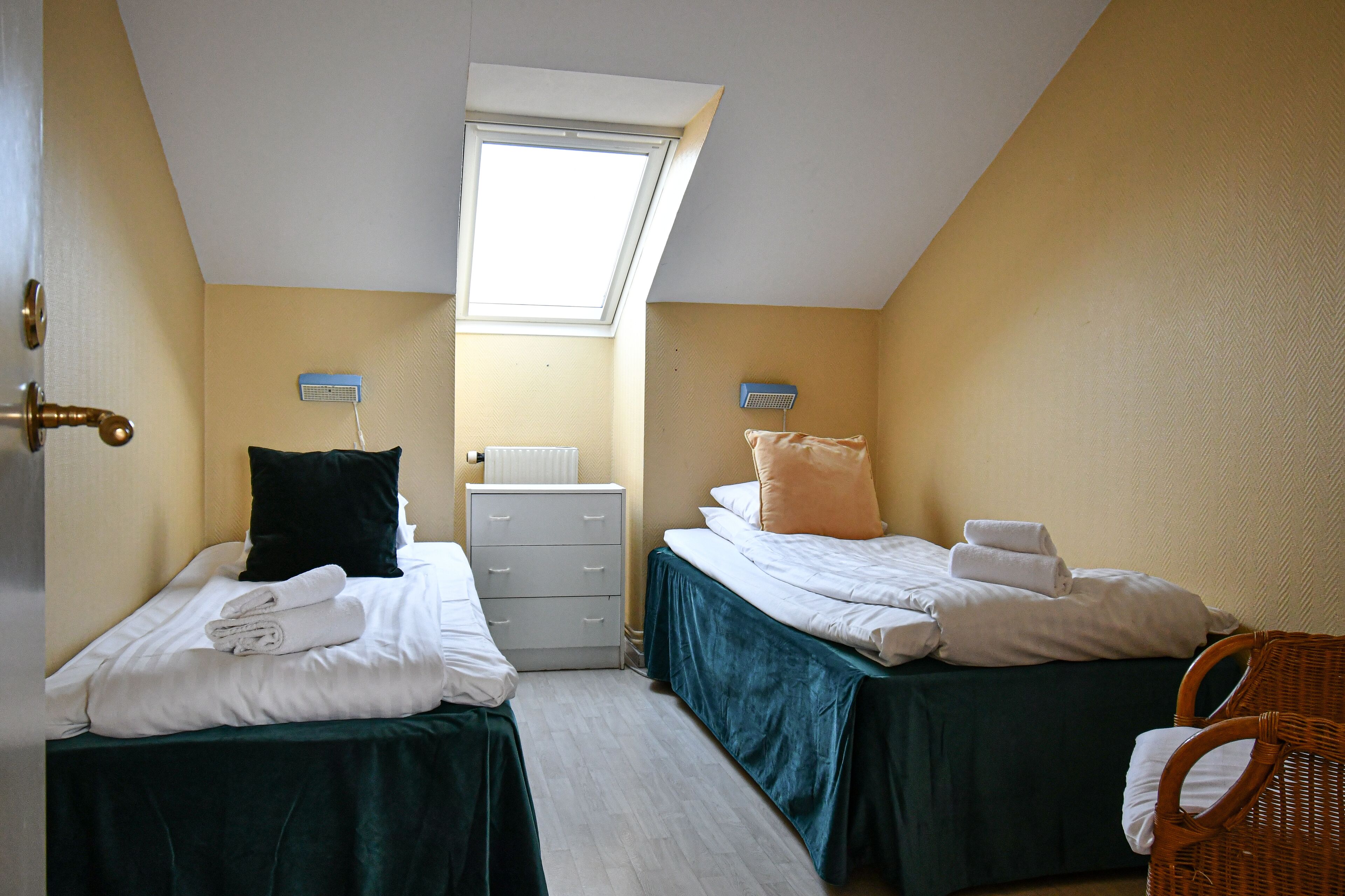 A room with two beds, a skylight, a white dresser, and a wicker chair.