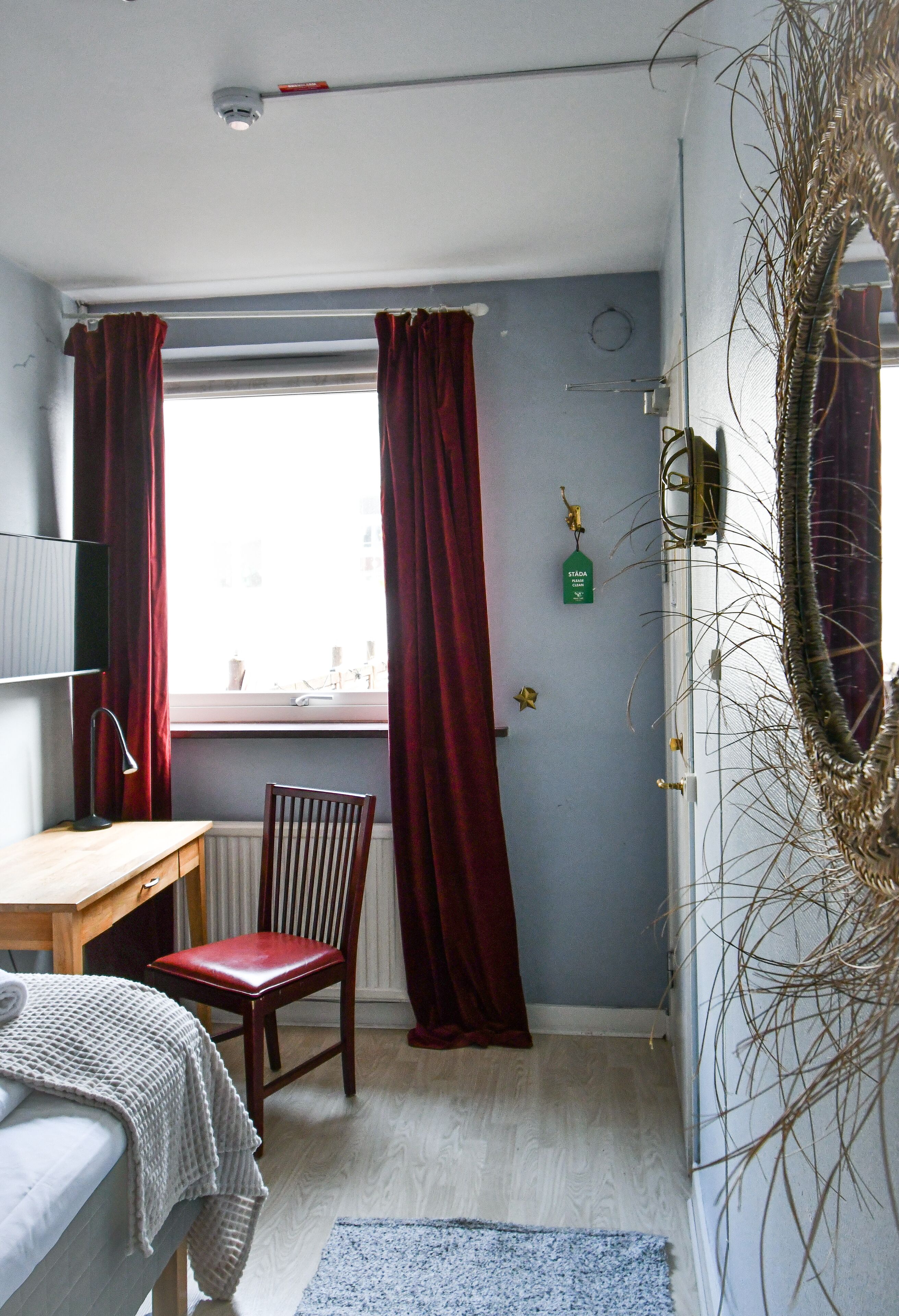 Een slaapkamer met een houten bureau, een stoel, een bed en een raam met rode gordijnen.