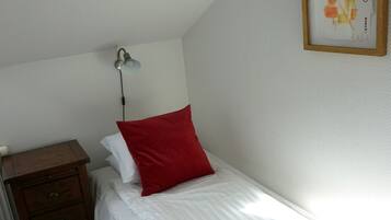 A neatly made bed with white linens and a red pillow, a bedside table with a lamp, and a framed artwork on the wall.
