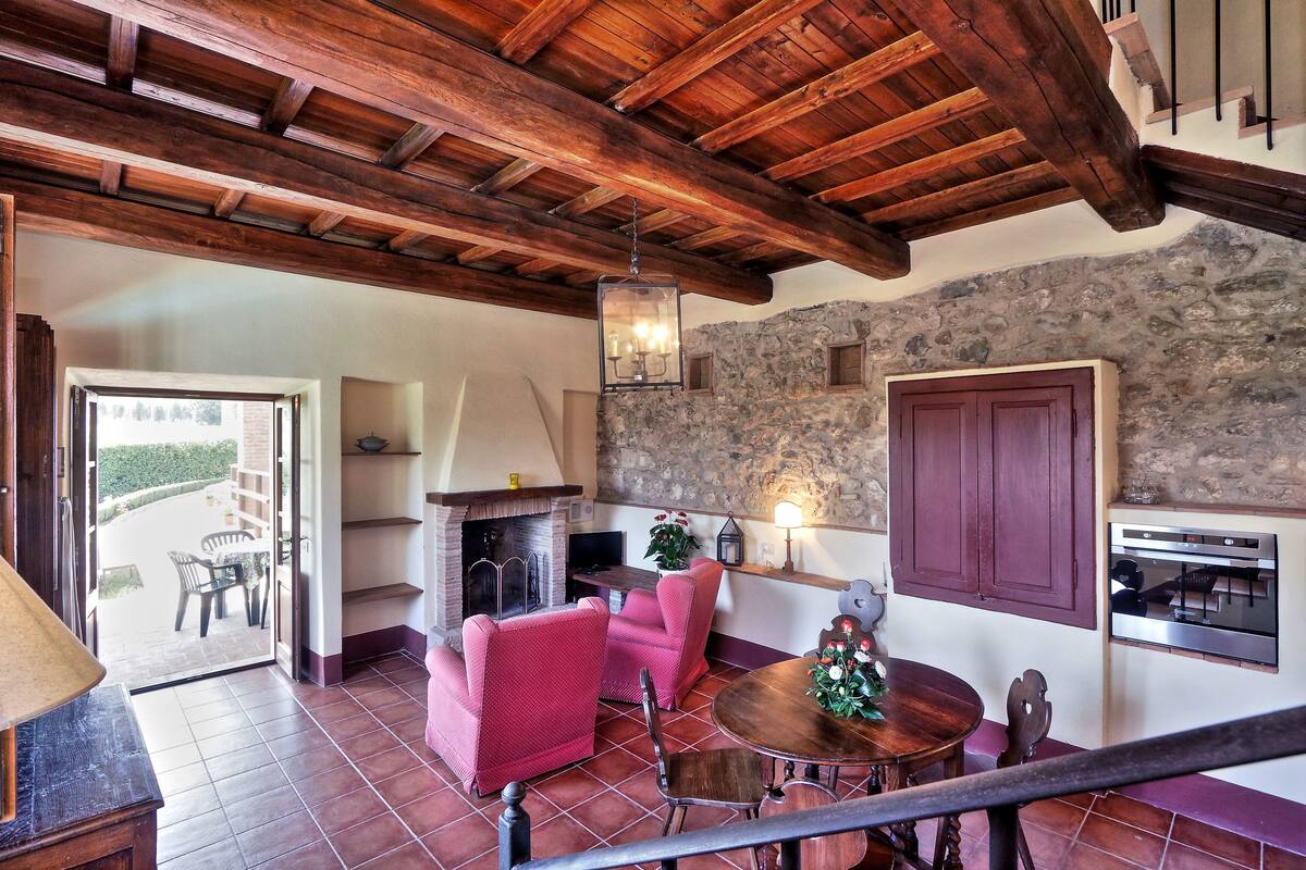 Une pièce chaleureuse avec un mur en pierre, des poutres apparentes en bois au plafond, une cheminée et un coin repas comprenant une table et des chaises.