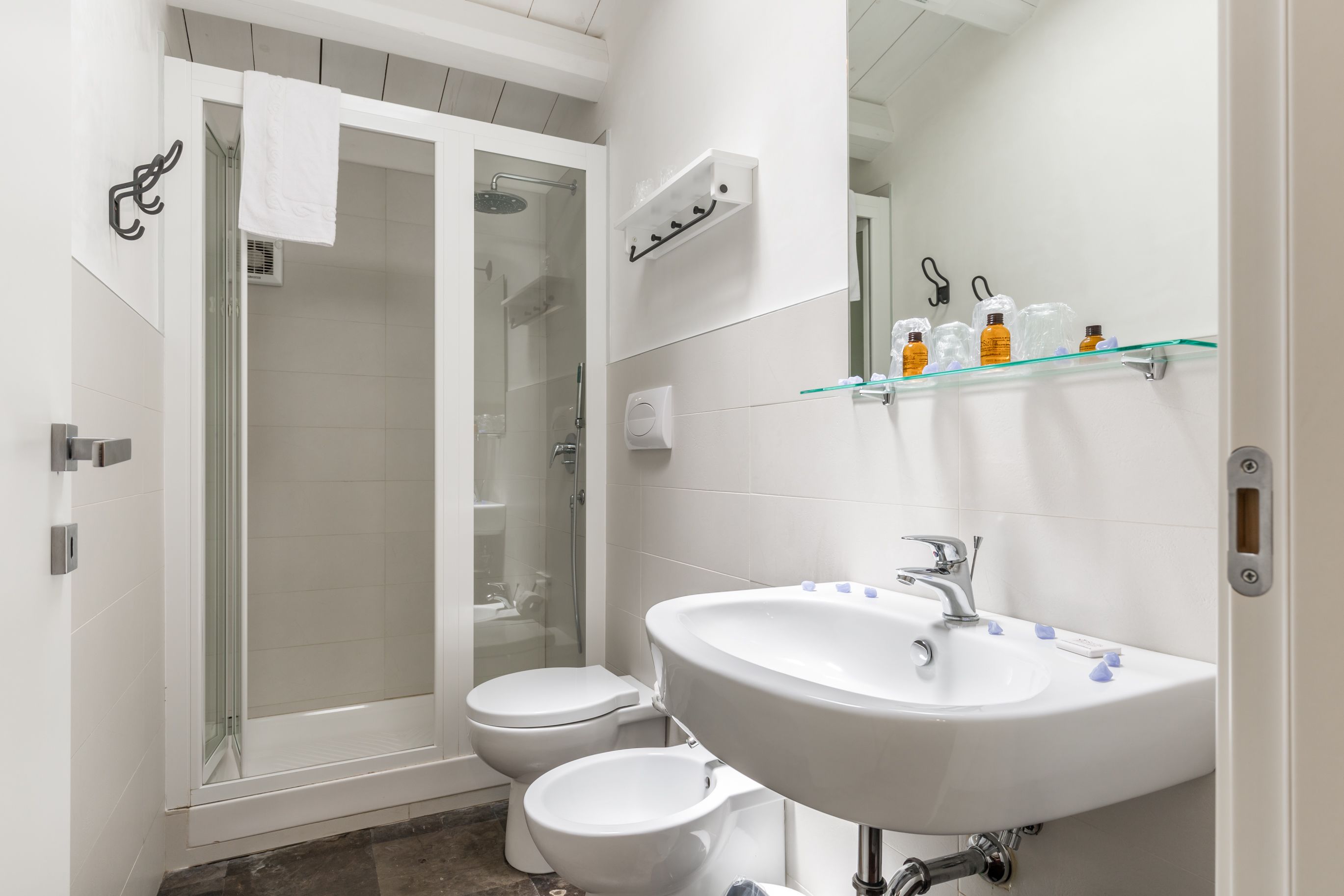 A modern bathroom with a glass-enclosed shower, two toilets, a large sink, and a glass shelf with toiletries.