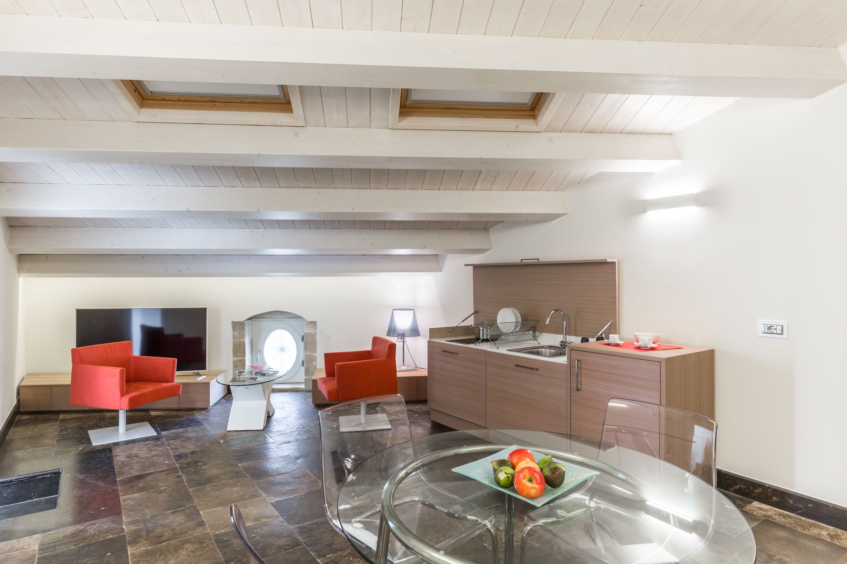 Un salon moderne avec une table en verre, des fauteuils rouges, un téléviseur à écran plat et un coin cuisine doté de meubles en bois et d’un évier.