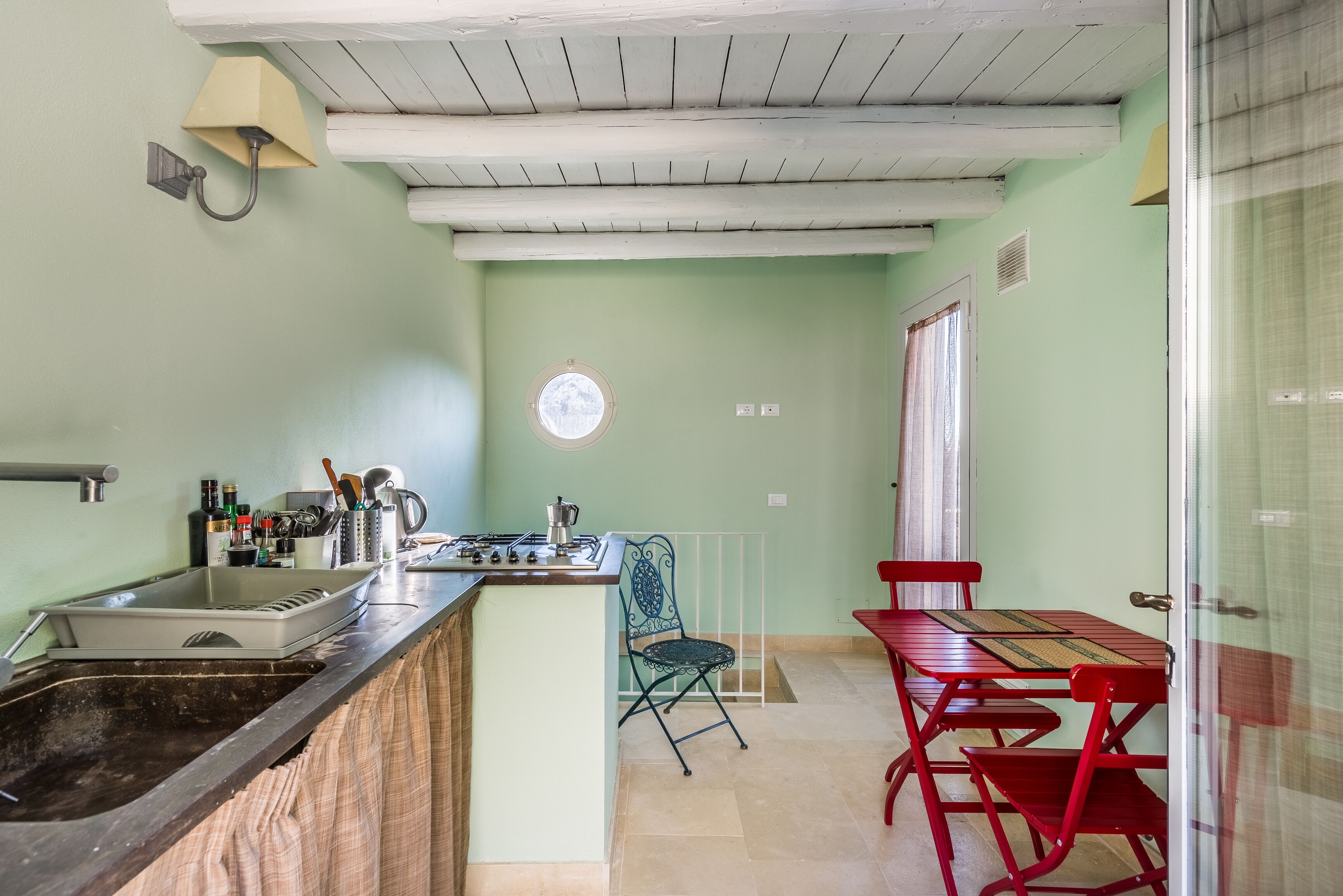 Een keuken met een spoelbak, fornuis en een raam. Er is een eethoek met een tafel en stoelen.