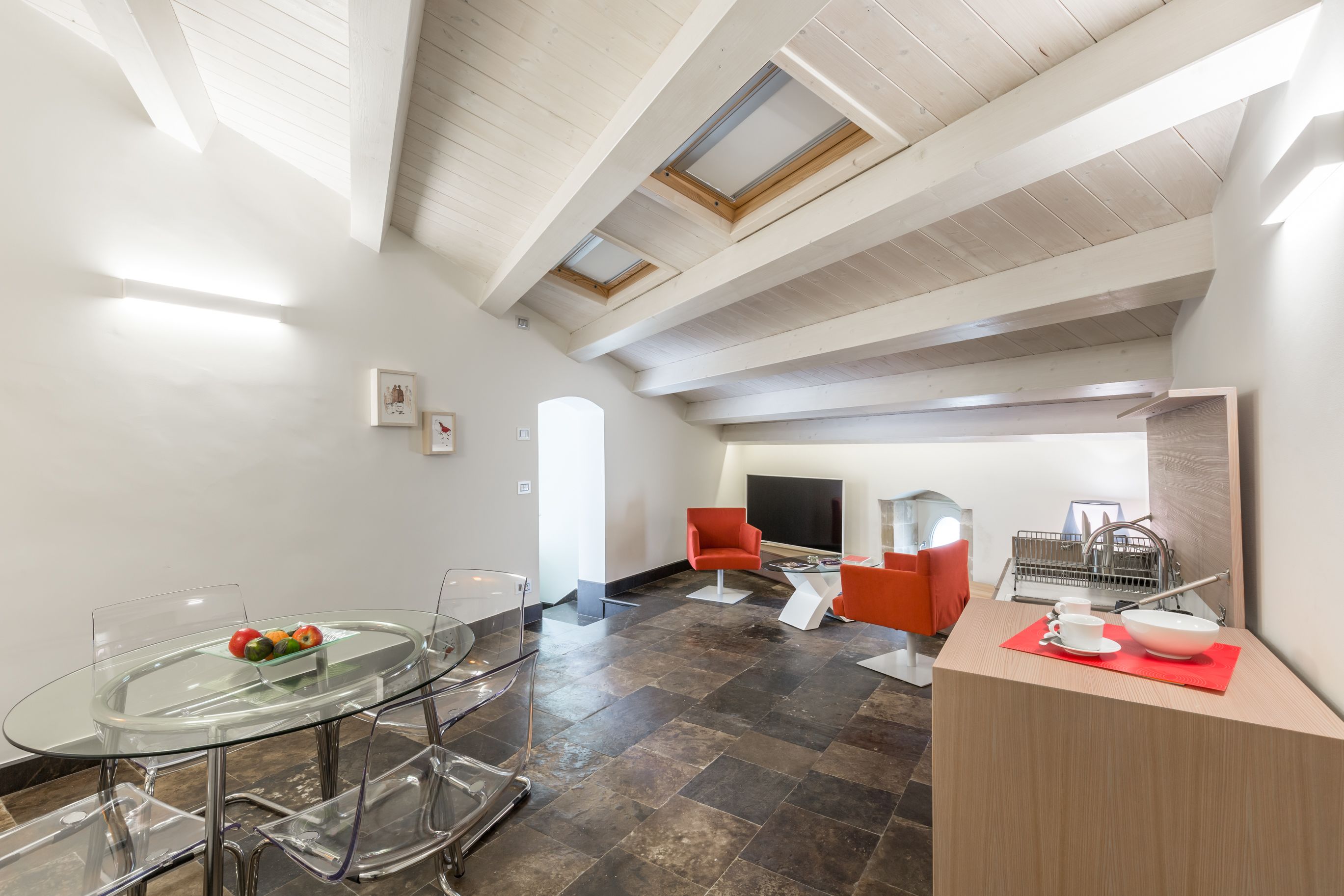 Um interior moderno com mesa de jantar de vidro, cadeiras vermelhas, área de cozinha e televisão.