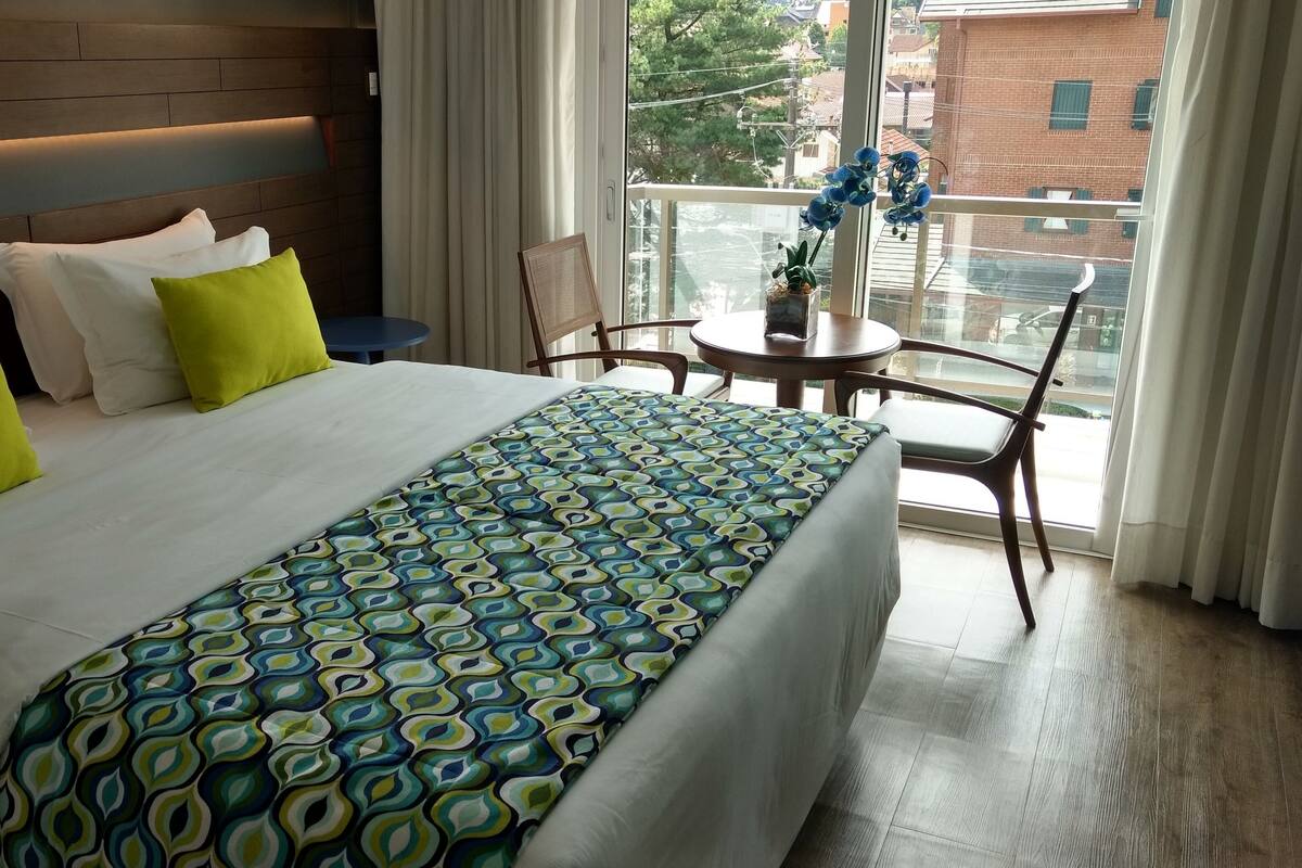 A hotel room with a large bed, a patterned bedspread, a small table with a plant, and a view of the outside through a window.