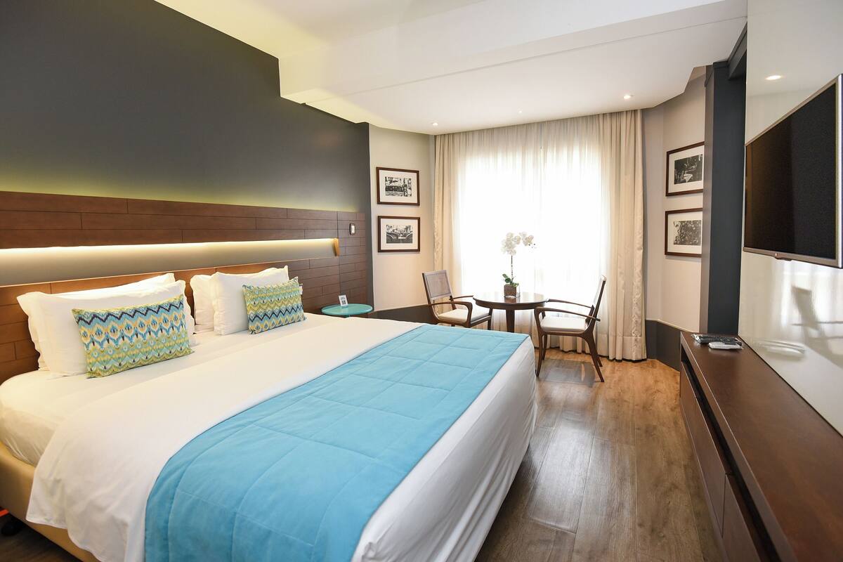 A hotel room with a large bed, a desk, a chair, a TV, and framed pictures on the wall.