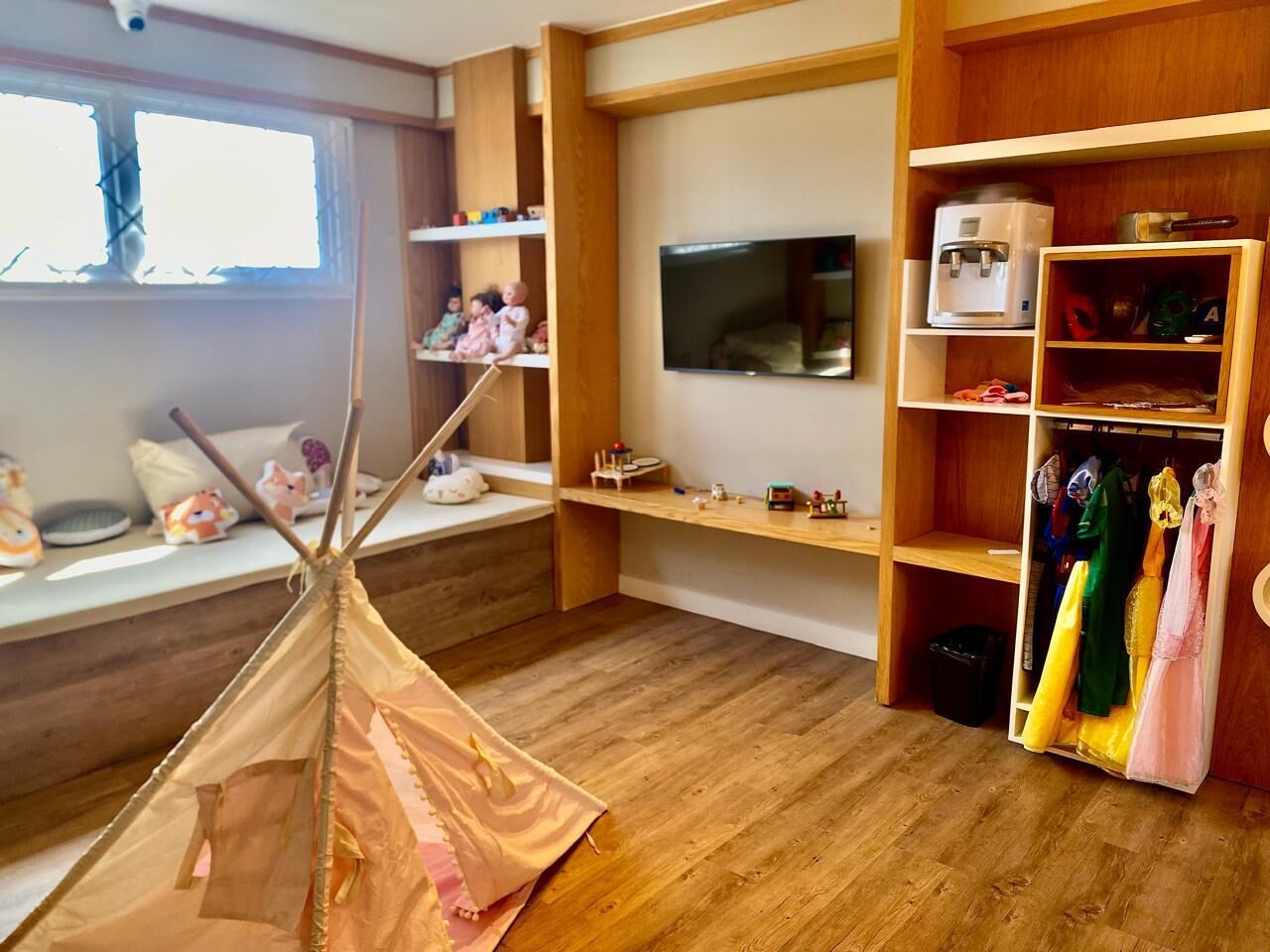 Habitación ideal para niños, con piso de madera, una carpa de juegos, televisor, estantes con juguetes y un perchero.