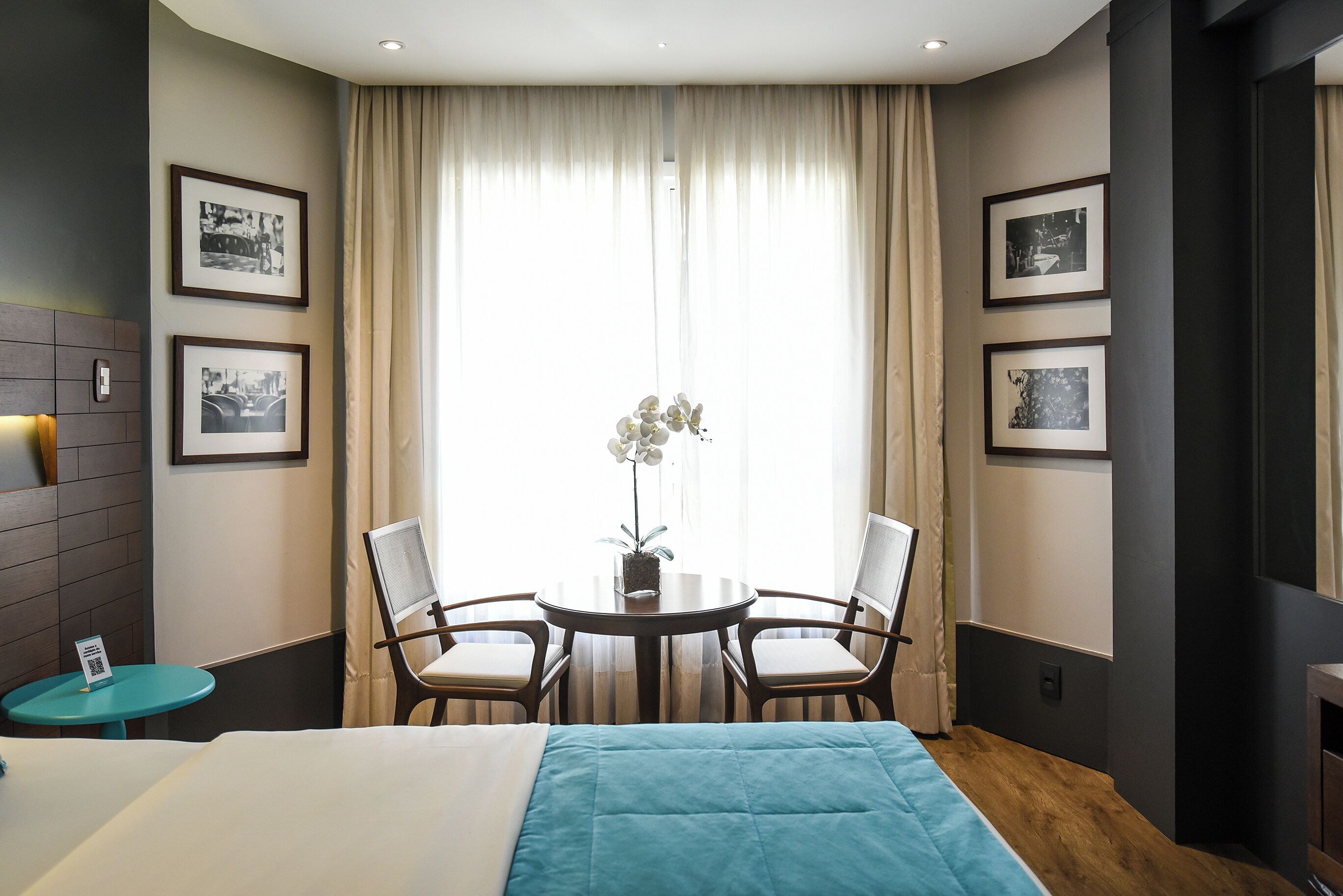 Habitación de hotel con una cama, una mesita redonda con sillas, una planta en maceta y cuadros en las paredes.