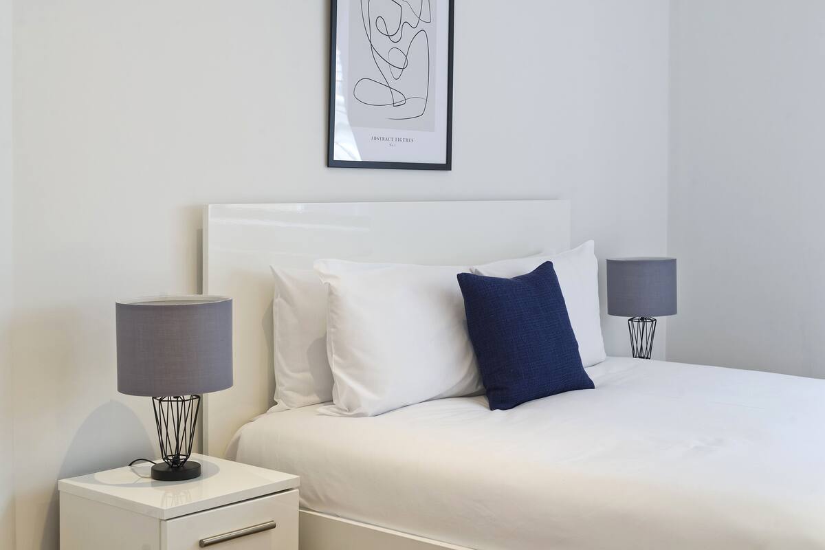 A neatly arranged bedroom with a white bed, white and blue pillows, a framed abstract artwork on the wall, and two bedside lamps on white nightstands.
