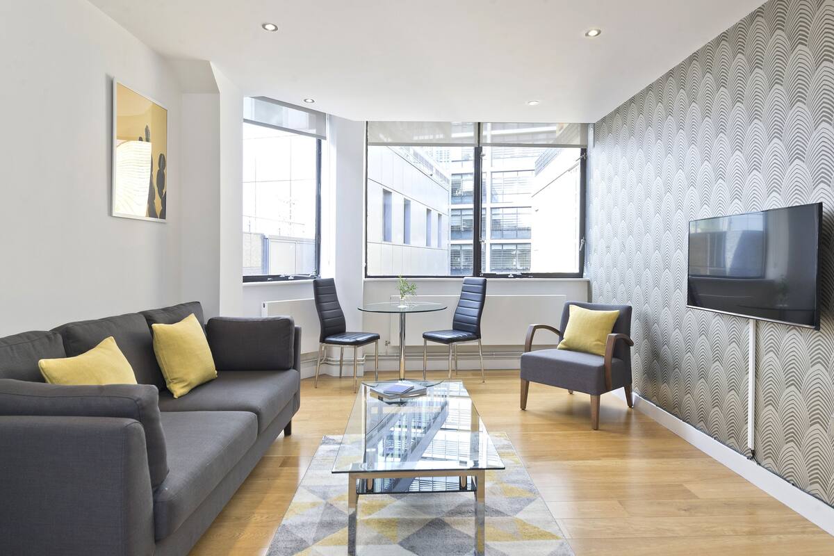 A modern living room with a grey sofa, two armchairs, a glass coffee table, and a TV mounted on the wall.