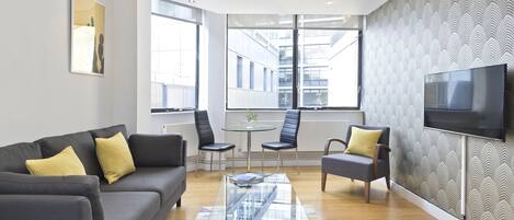Un salon moderne avec un canapé gris, deux fauteuils, une table basse en verre et un téléviseur fixé au mur.