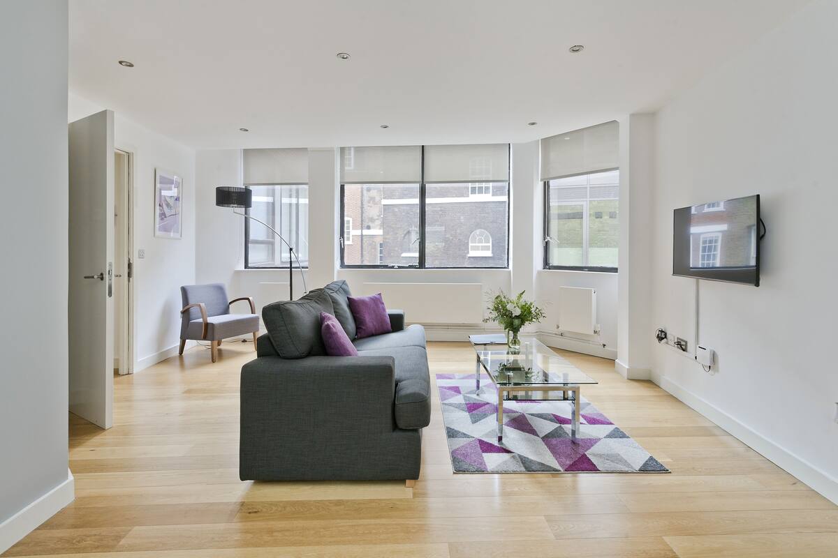 A modern living room with a grey sofa, a glass coffee table, and a view of a brick building through large windows.
