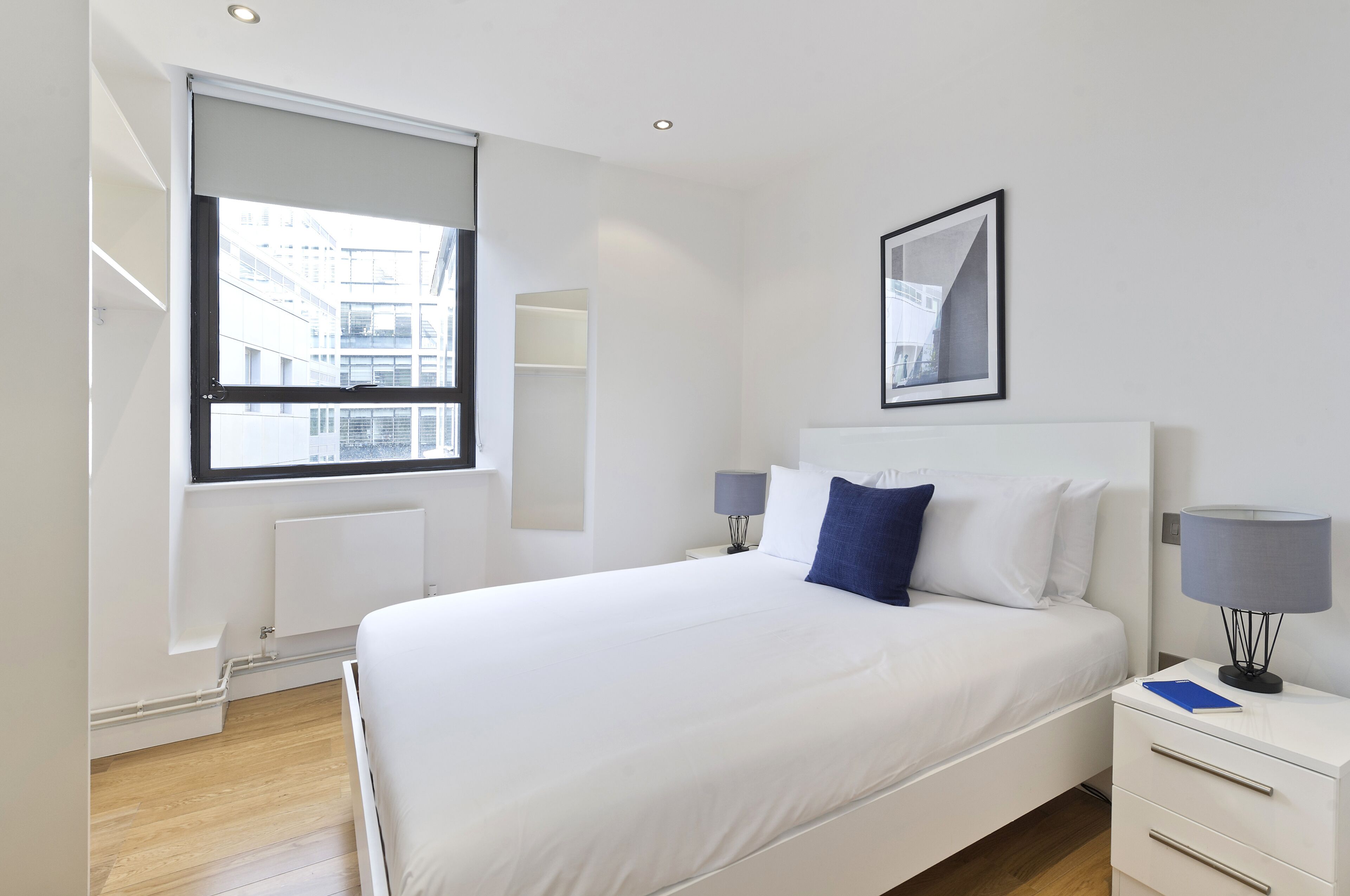 A modern hotel room with a large bed, a nightstand, a window with a view of buildings, and a framed picture on the wall.