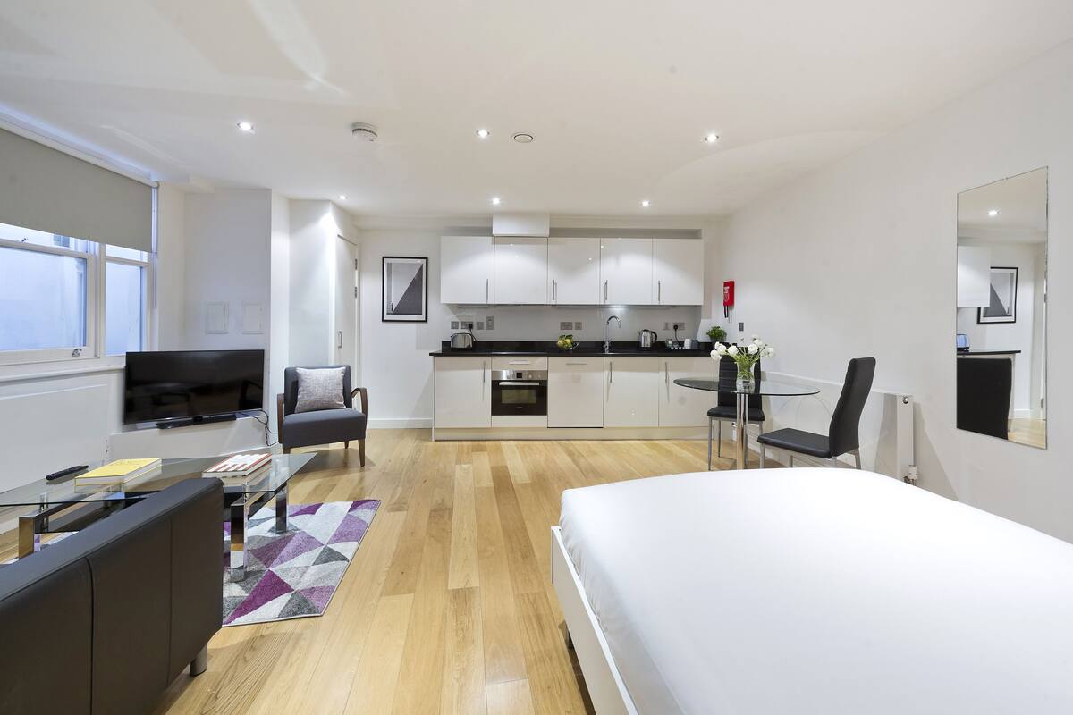 A modern hotel room with a large bed, a glass coffee table, a sofa, a TV, a kitchen area with cabinets and a sink, and a small dining table with chairs.