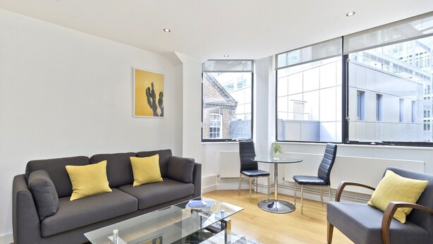A modern living room with a grey sofa, a glass coffee table, and a yellow cushion. There is a dining area with a small table and chairs, and large windows offering a view of the city. - Red Lion Court by City2Stay (London)