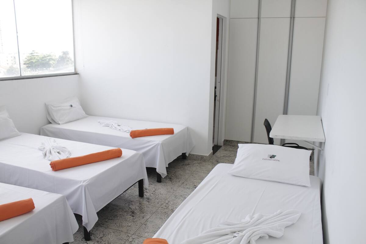 A hotel room with three single beds, each with white linens and orange accents. There is a small table with a lamp and a chair.