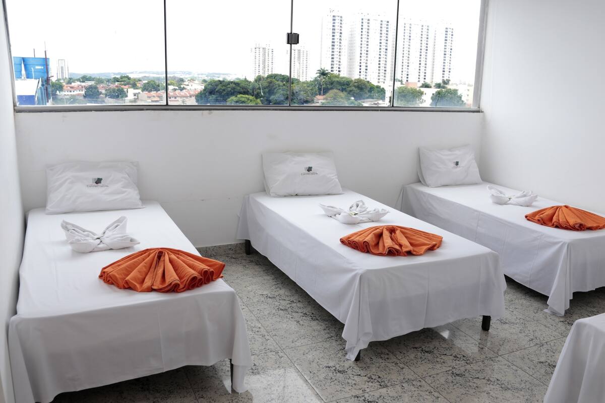 A hotel room with three single beds, each with white bedding and orange towels. The room has a large window offering a city view.