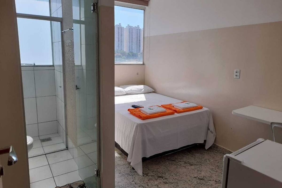 A hotel room with a bed, a small table, a shower area, and a view of buildings outside the window.