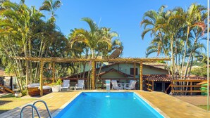 A rectangular swimming pool with a wooden deck surrounding it, a pergola with white chairs, and palm trees in the background. - Pousada Sonho de Geribá (Búzios)