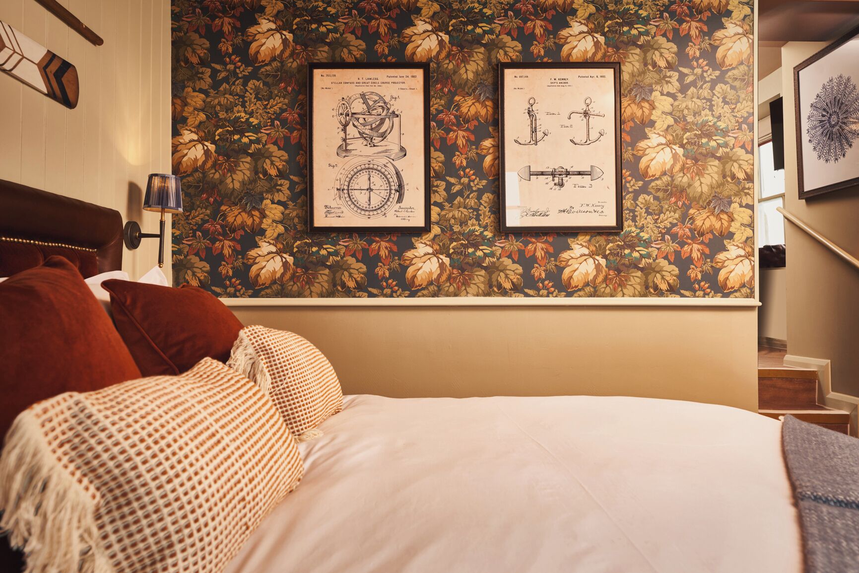 Un dormitorio con una cama, almohadas, una lámpara fijada a la pared y dos dibujos técnicos enmarcados.