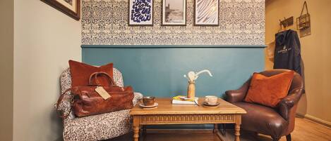 A room with a patterned wallpaper, a wooden coffee table, a leather armchair, and a sofa with a floral cushion.