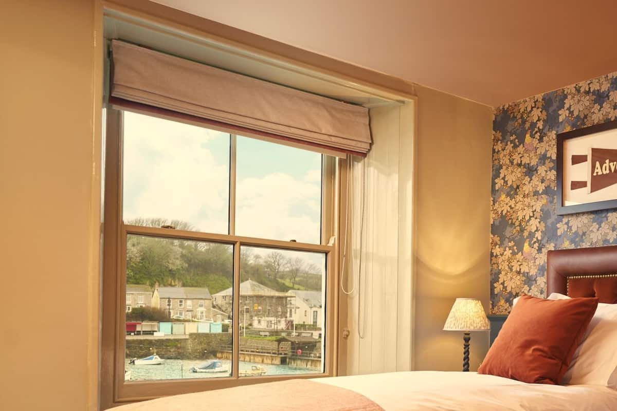 A hotel room with a large window, a bed with a headboard, a lamp, and a floral wallpaper.