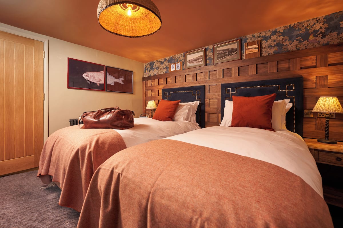 A hotel room with two beds, wooden headboards, a wooden wardrobe, and a wall decorated with framed pictures.