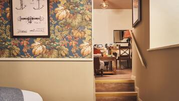 Un escalier menant à une salle à manger dont les murs sont recouverts d’un papier peint à motifs floraux et où est accroché un plan technique encadré.