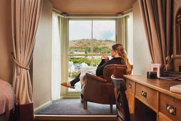 A hotel room with a large window, a wooden desk, a chair, and a view of a landscape. - Harbour Inn (Helston)