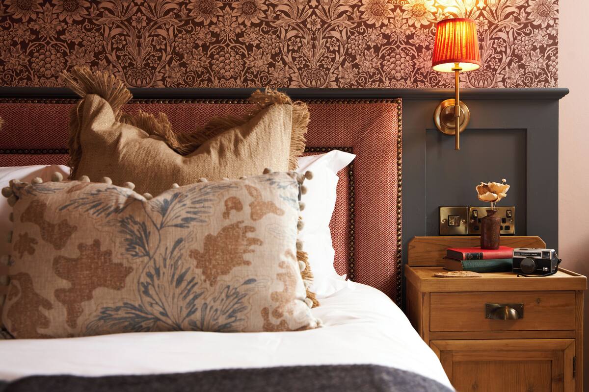 A neatly made bed with patterned pillows, a wooden nightstand with books and a camera, and a wall with floral wallpaper.