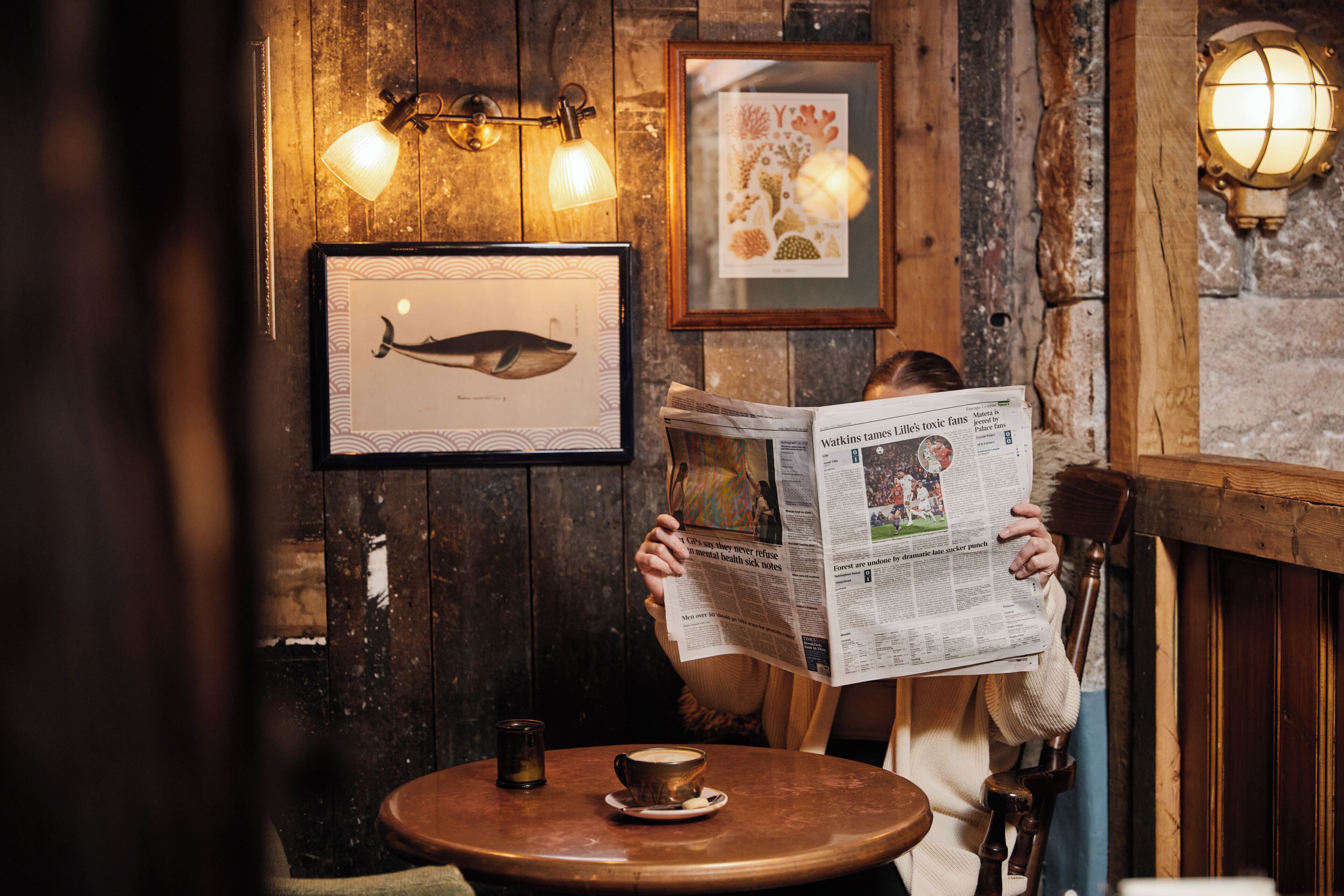 A person reading a newspaper at a wooden table with a cup and saucer, in a room with wooden walls and framed artwork.