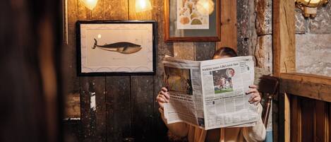 A person reading a newspaper at a wooden table with a cup and saucer, in a room with wooden walls and framed artwork.