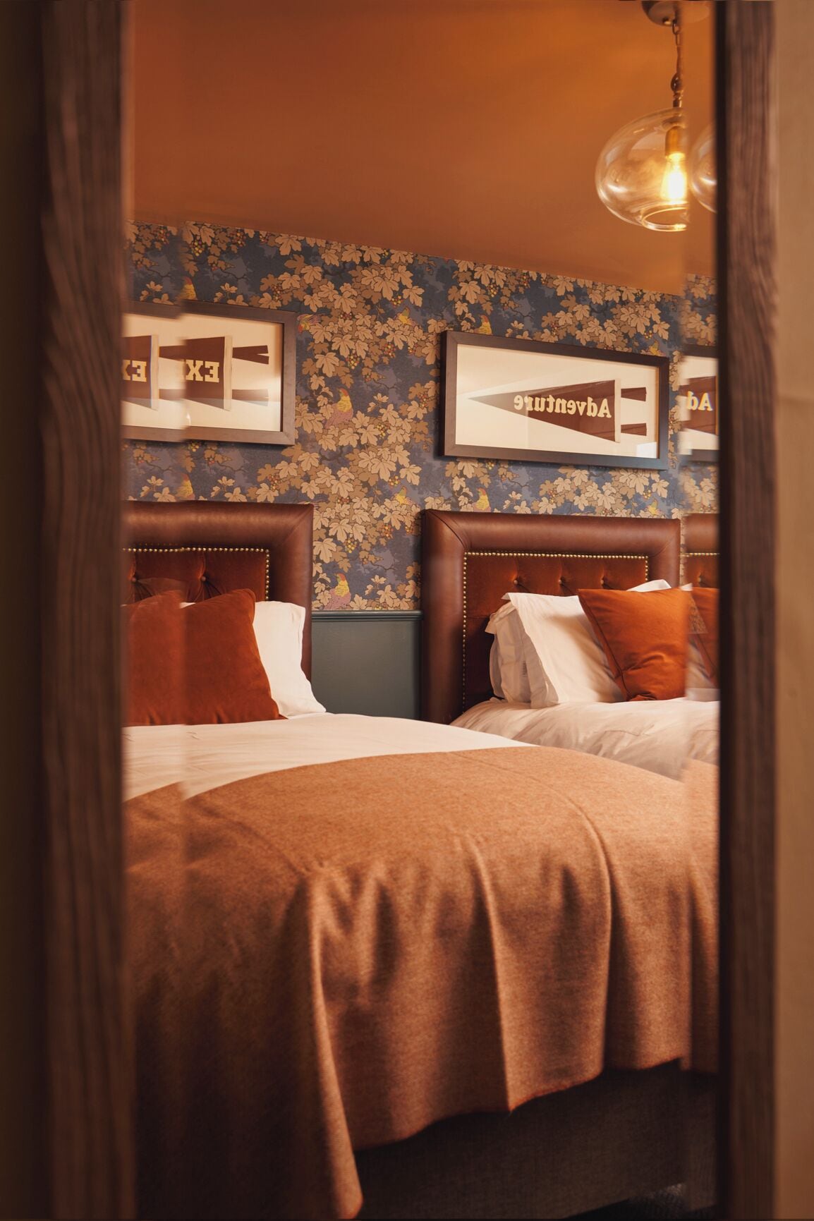 Habitación de hotel con dos camas, papel tapiz floral, cuadros enmarcados y una lámpara colgante.