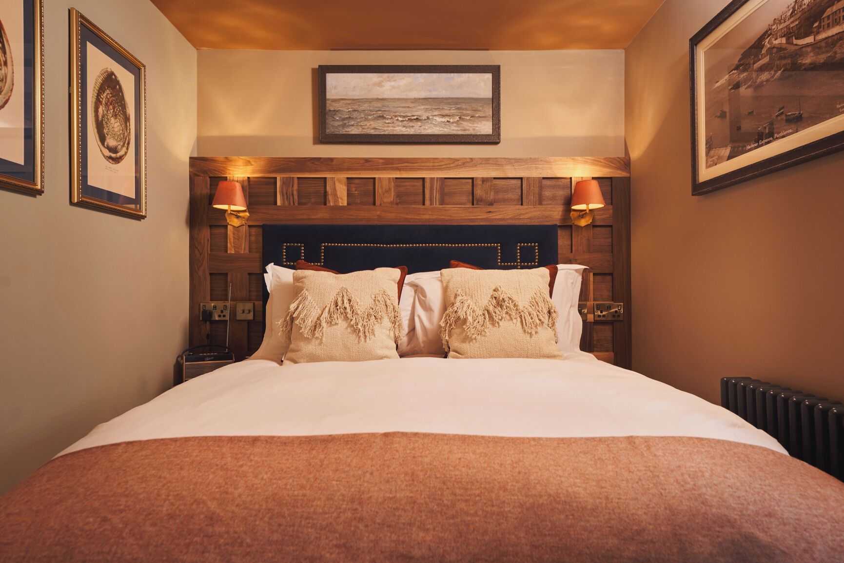 A hotel room with a wooden headboard, two bedside lamps, a framed picture above the bed, and a bed with a beige throw blanket.