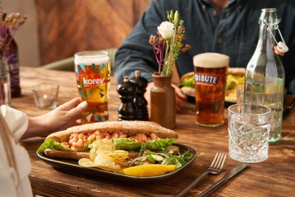 A dining table with a sandwich, a beer glass, a bottle, and a glass of water.