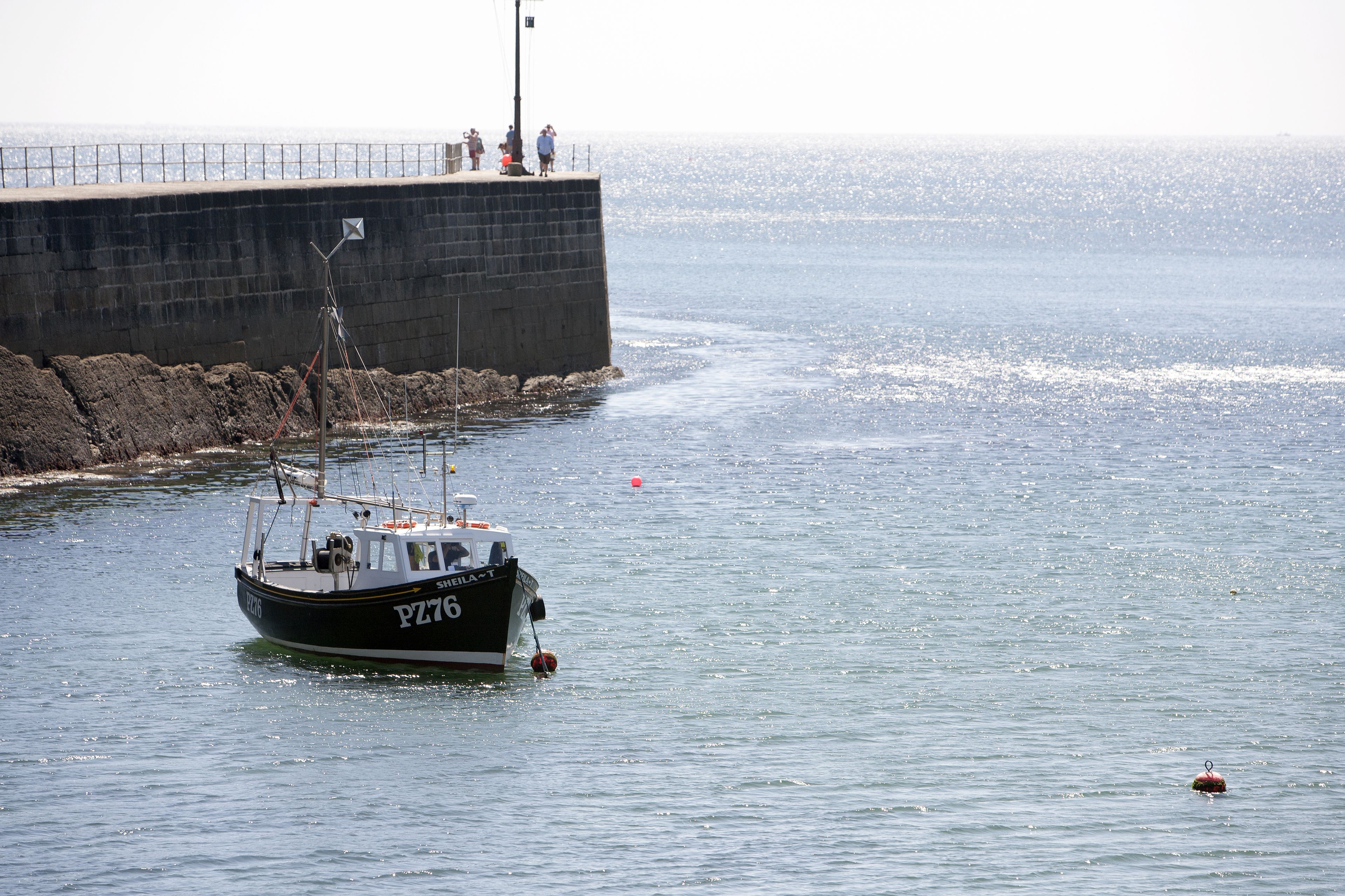 A boat named "PZ76" anchored near a pier with a rocky breakwater and a clear view of the sea.