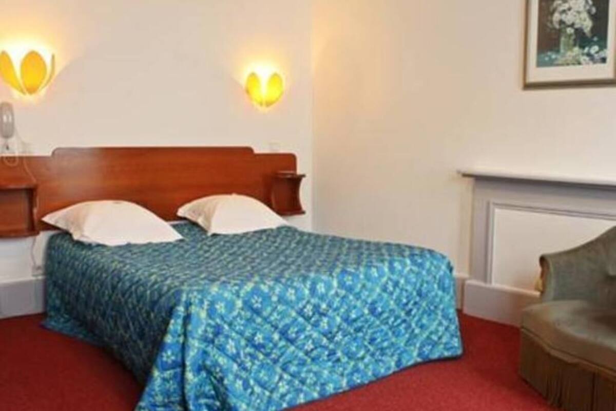 Une chambre d’hôtel avec une tête de lit en bois, une couvre-lit bleue à motifs, deux lampes fixées au mur et une moquette rouge.