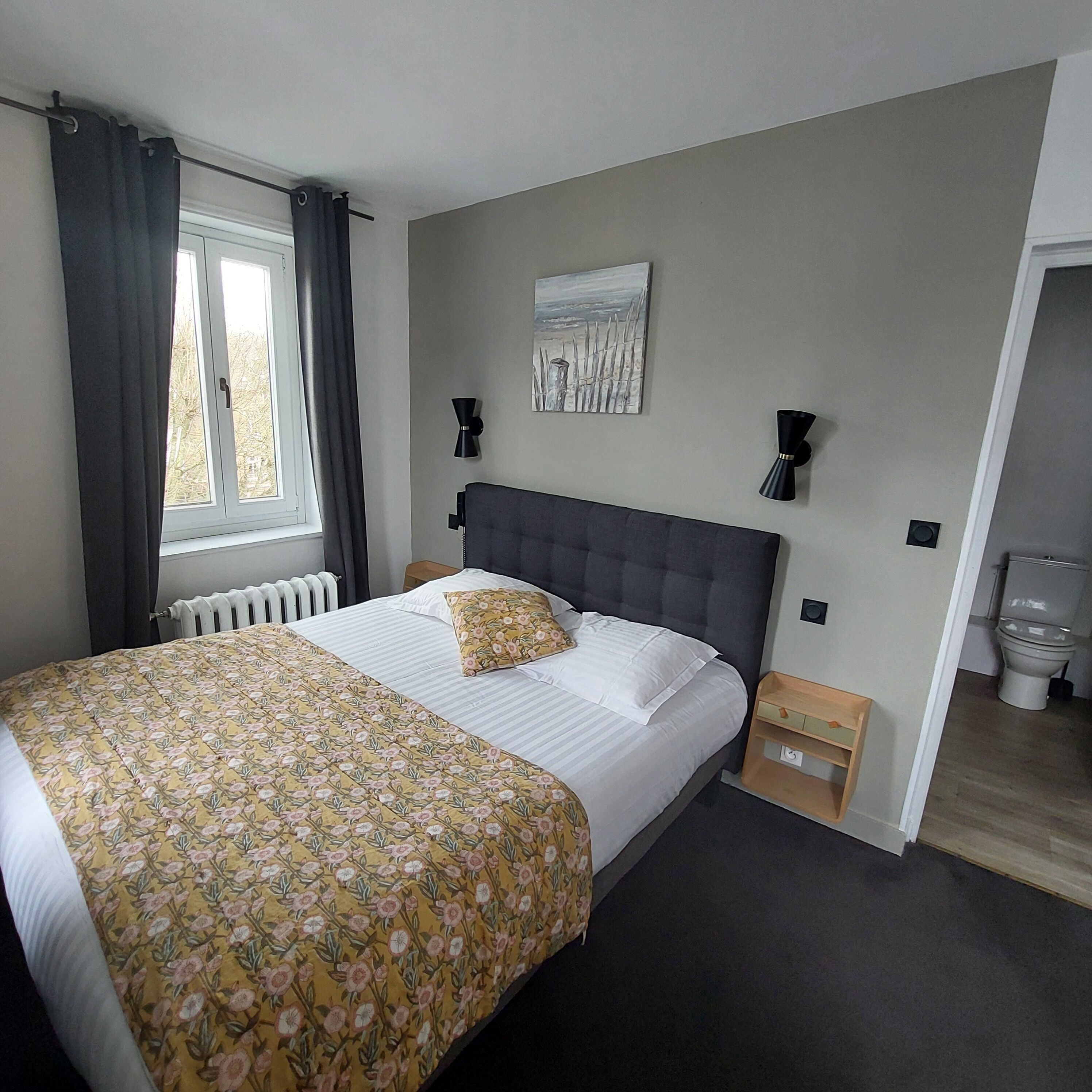 Un dormitorio con cama, radiador, ventana con cortinas, un cuadro en la pared y una mesita de noche de madera.