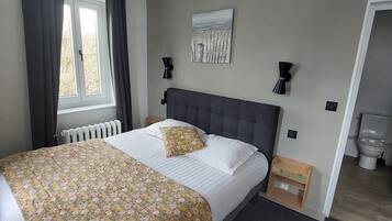 Un dormitorio con cama, radiador, ventana con cortinas, un cuadro en la pared y una mesita de noche de madera.