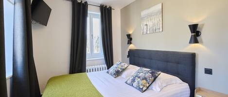 Un lit bien fait, avec une couverture verte et des coussins à motifs floraux, un téléviseur fixé au mur et deux appliques murales.