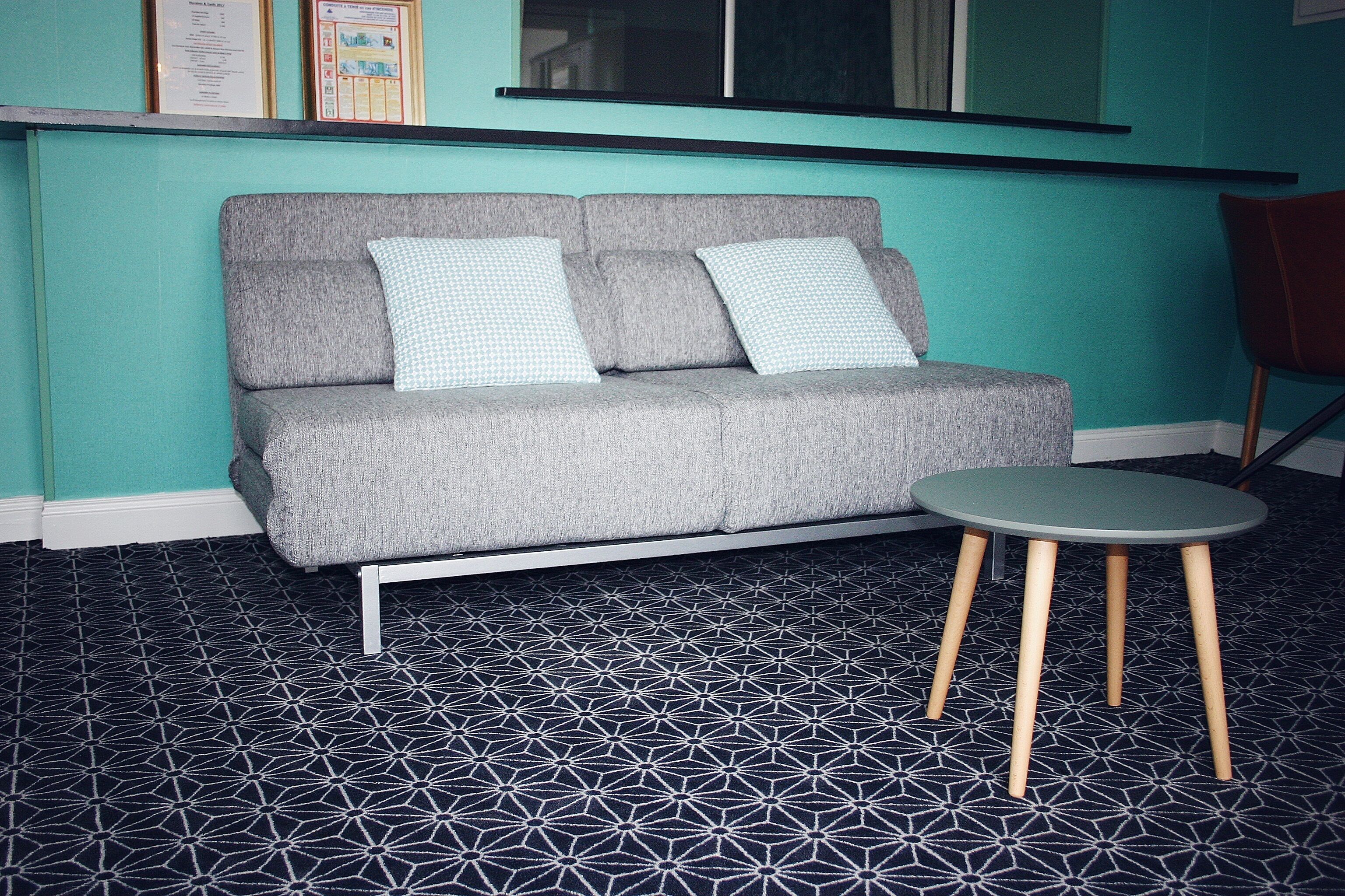 A grey sofa with two patterned cushions, a small round table with wooden legs, and a geometric patterned floor.