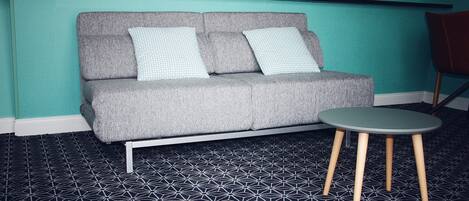 A grey sofa with two patterned cushions, a small round table with wooden legs, and a geometric patterned floor.