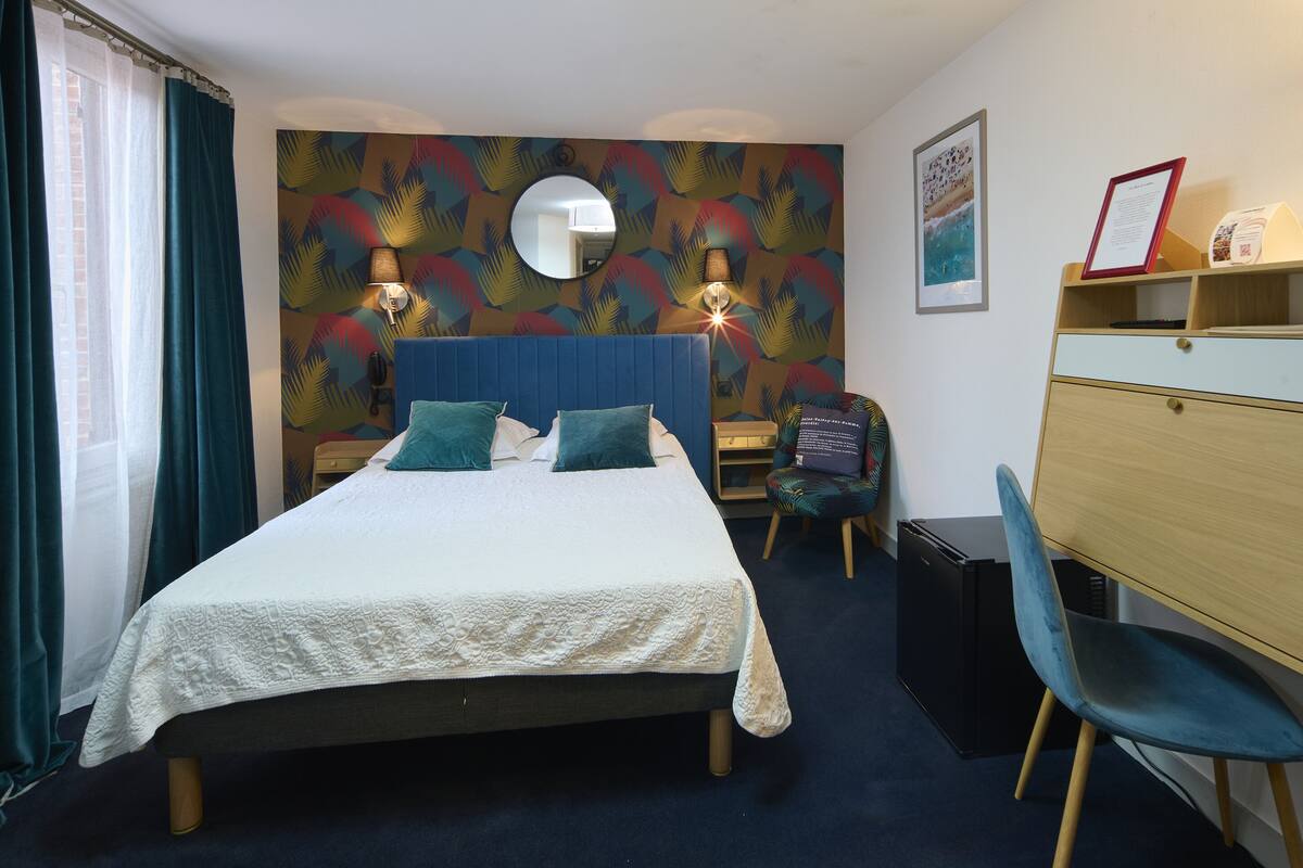 Une chambre d’hôtel comprenant un lit, un bureau, une chaise, une lampe, un miroir et un mur orné d’un motif de feuilles tropicales.